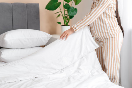 Young woman in pajamas making bed at home