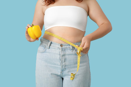 Overweight young woman with measuring tape and bell pepper on blue background, closeup. Weight gain conceptの写真素材