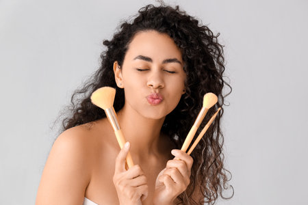 Beautiful young African-American woman with brush makeupes blowing kiss on gray backgroundの写真素材
