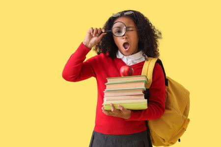 Shocked little African-American schoolgirl with magnifier and books on yellow backgroundの写真素材