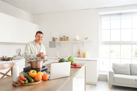Young man with laptop and fresh vegetables writing recipe in kitchenの写真素材