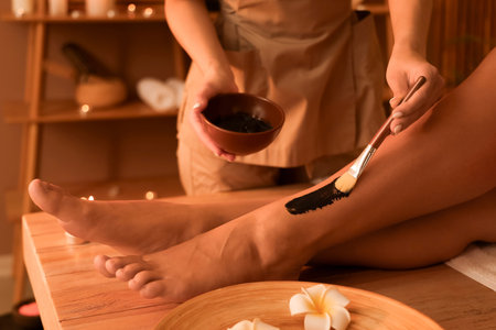Massage therapist applying mud on woman's leg in dark spa salon, closeupの写真素材