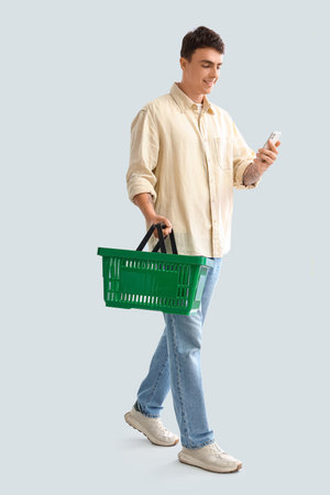Young man with shopping basket and mobile phone walking on light backgroundの写真素材