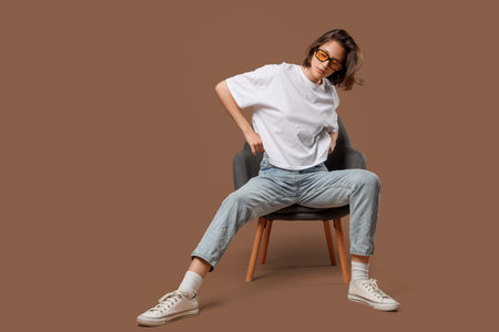 Beautiful young woman in stylish jeans sitting on chair against brown backgroundの写真素材