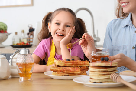 Cute little girl and her mother pouring honey on waffles in kitchen, closeupの写真素材