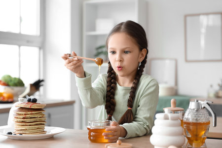 Cute little girl with honey and pancakes at table in kitchenの写真素材