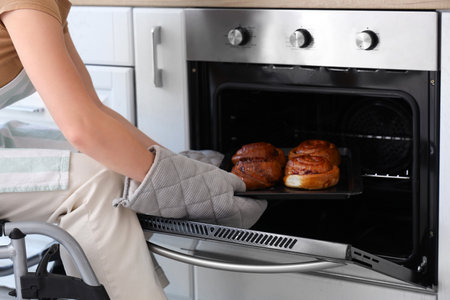 Young woman in wheelchair taking baking tray with buns from oven at home, closeupの写真素材