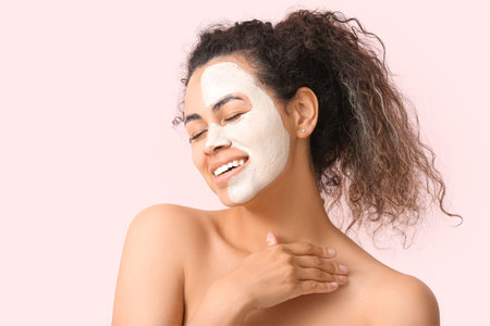 Happy young African-American woman with facial mask on pink background