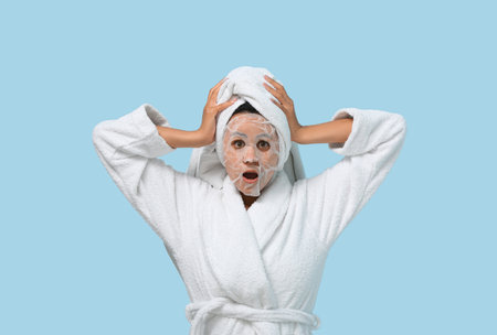 Surprised young African-American woman in bathrobe with sheet facial mask on blue backgroundの写真素材