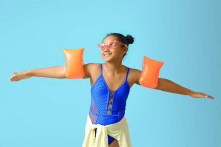 Happy African-American girl in swimsuit with inflatable armbands on blue background