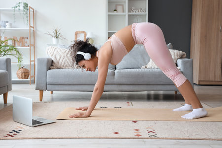 Sporty African-American woman with laptop and headphones exercising on yoga mat at homeの写真素材