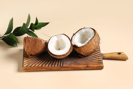 Wooden board with halves of fresh coconuts and leaves on beige background