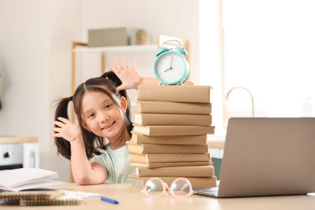Funny little Asian girl with books and alarm clock studying online in kitchenの写真素材