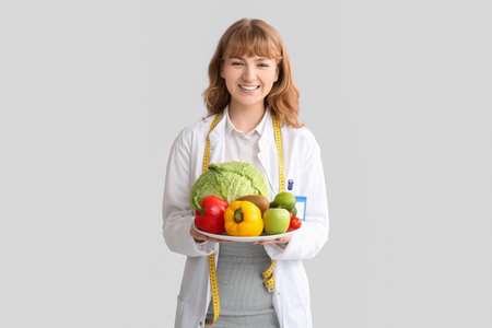 Beautiful young happy female nutritionist with fresh vegetables and measuring tape on gray backgroundの写真素材