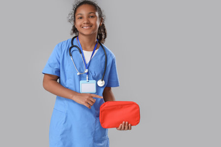 Little African-American doctor pointing at first aid kit on gray backgroundの写真素材