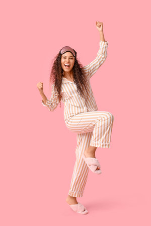 Beautiful young happy African-American woman in pajamas with mask on pink background