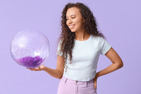 Beautiful young African-American woman with beach ball on lilac backgroundの写真素材