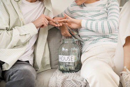Young couple putting money into jar on sofa at home, closeup