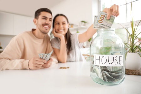 Young couple putting money into jar at table in kitchen