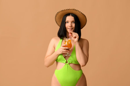 Beautiful young woman in stylish green swimsuit with glass of cocktail on brown backgroundの写真素材