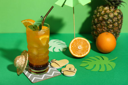 Glass of tasty cocktail with orange, pineapple, paper palm leaves and flip flops on green background