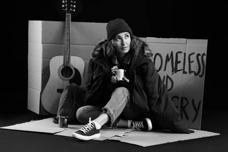 Mature homeless woman with can sitting on cardboard against dark background