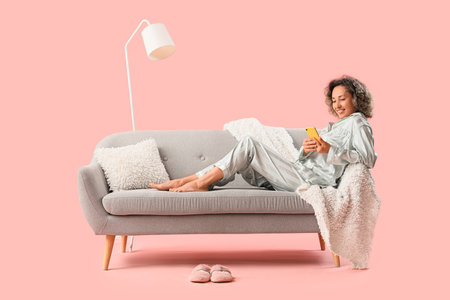 Mature woman in pajamas using mobile phone on gray sofa against pink backgroundの写真素材