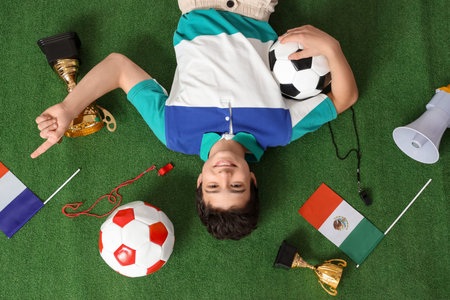 Teenage boy with soccer balls, flags and gold cups lying on green grass