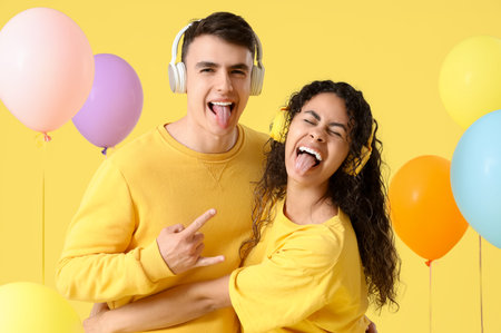 Young couple in love with headphones and balloons showing tongues on yellow backgroundの写真素材
