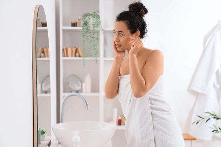 Young African-American woman removing makeup with cotton bud in bathroomの写真素材