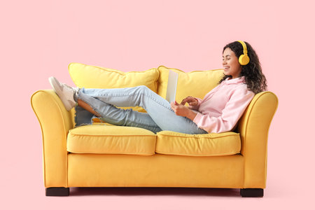 Young African-American woman with headphones using laptop on yellow sofa against pink backgroundの写真素材