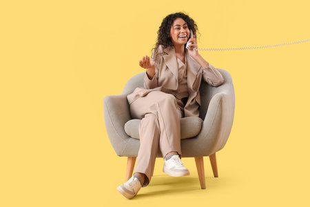 Young African-American businesswoman talking by phone in gray armchair on yellow backgroundの写真素材