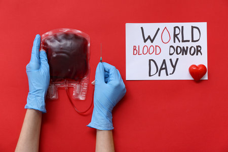Female hands in rubber gloves with blood pack and paper sheet with text WORLD BLOOD DONOR DAY on red backgroundの写真素材