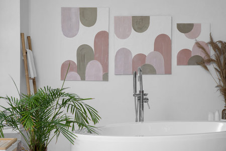 Stylish bathroom with bathtub, houseplant and pictures on white wall