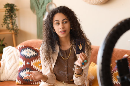 Young African-American witch with tarot card recording video at homeの写真素材