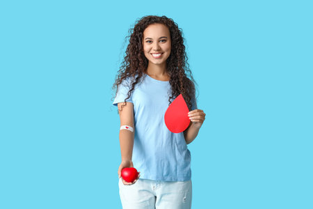 Young female African-American donor with applied medical patch, paper blood drop and heart on blue backgroundの写真素材