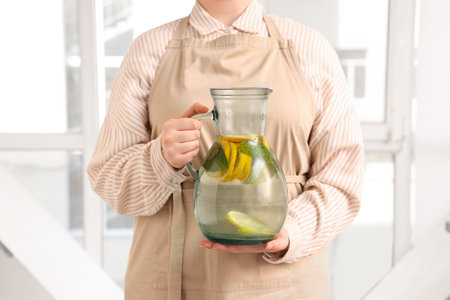 Young woman holding jug of lemon infused water with mint near window at homeの写真素材