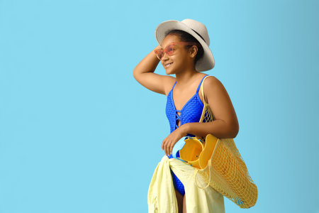 Happy African-American girl in swimsuit with string bag and beach accessories on blue backgroundの写真素材