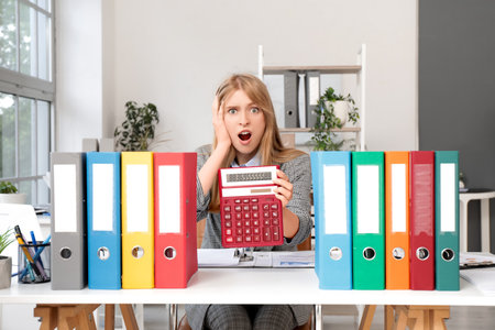 Shocked young female accountant showing calculator at workplace with stacks of folders in officeの写真素材