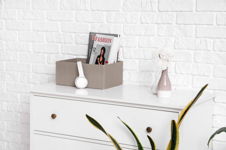 Chest of drawers with magazines, headphones, flowers and hourglass near white brick wall