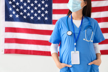 Female doctor in medical mask against USA flag on light background. Labor Day celebrationの写真素材