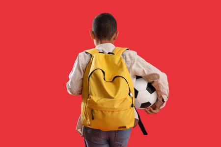 Little schoolboy with backpack and soccer ball on red background, back viewの写真素材