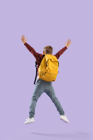 Little schoolboy with yellow backpack jumping on lilac background, back viewの写真素材