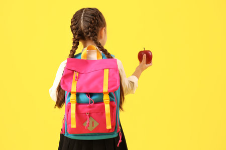 Little schoolgirl with backpack and apple on yellow background, back viewの写真素材