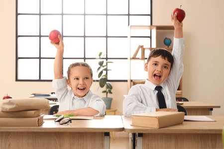 Happy little schoolchildren with apples at desks in classroom