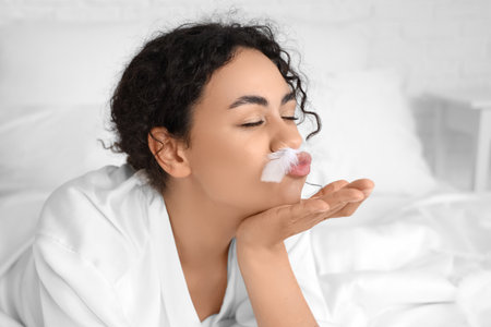 Young African-American woman with feather blowing kiss in bedroom, closeupの写真素材