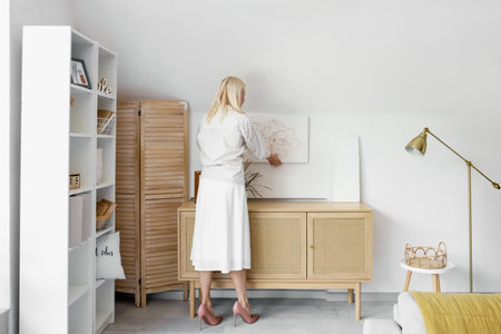 Mature woman hanging beautiful painting on light wall at home, back viewの写真素材