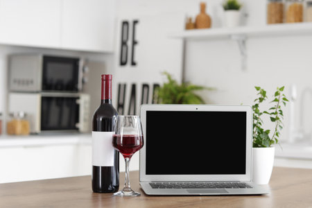 Bottle, glass of wine and laptop on wooden table in kitchen, closeupの写真素材