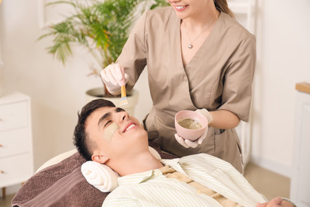 Beautician applying clay mask on young man's face in salonの写真素材