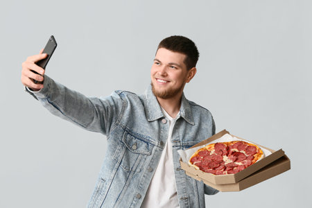 Young man holding cardboard box with tasty pepperoni pizza and taking selfie on gray backgroundの写真素材